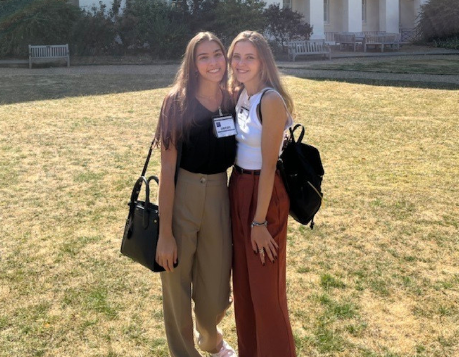 No centro da imagem, duas amigas se abraçam. Elas vestem calça e blusas de manga curta. Ambas têm mochilas. Ao fundo, é possível ver um gramado que se estende em direção a um prédio. O dia é claro e ilumina a imagem.